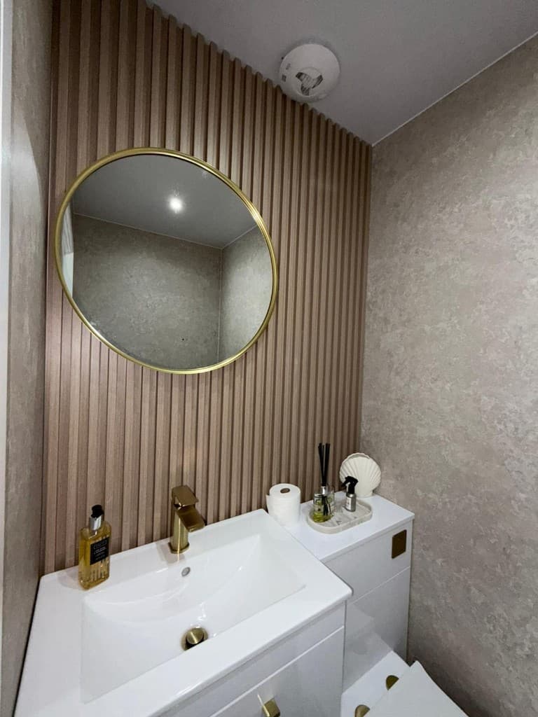 Stylish bathroom with circular gold mirror, vertical wood slats, and sanitised white vanity after guest turnover