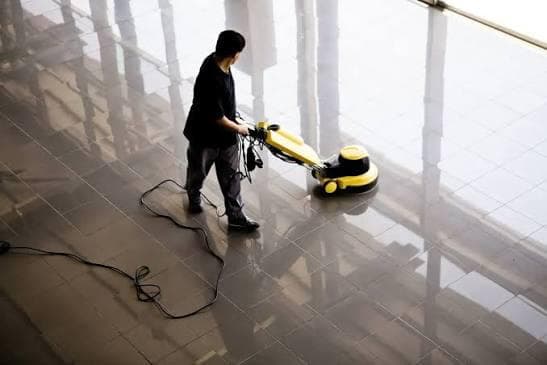Cleaner operating a yellow industrial floor buffer on reflective tiled flooring in a spacious commercial premises