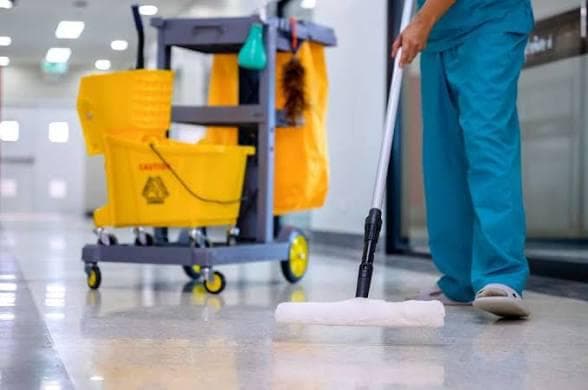 Professional cleaner in blue scrubs mopping a polished corridor floor with a janitorial trolley in a bright commercial building