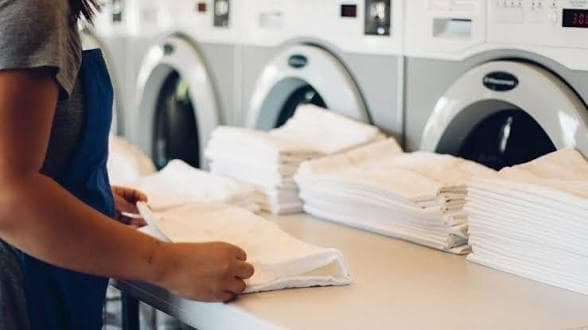 Laundry operative folding clean white linens at a counter with industrial washing machines in a commercial laundry