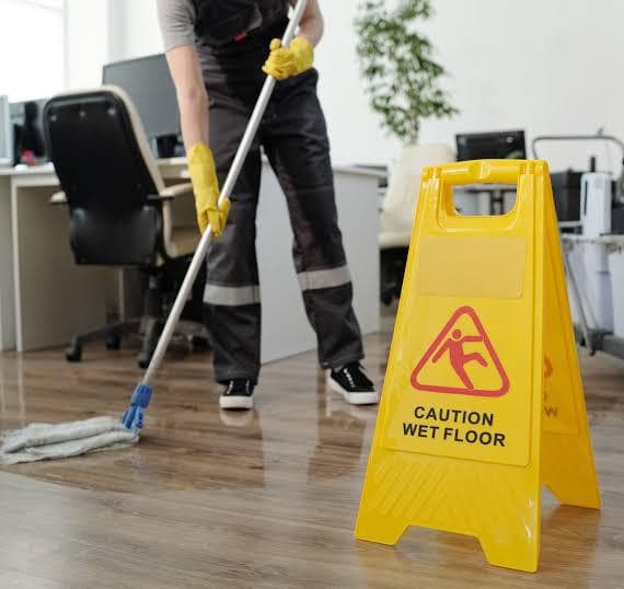 Professional cleaner wearing yellow gloves mopping a polished office floor with a wet-floor safety sign visible in a modern workspace