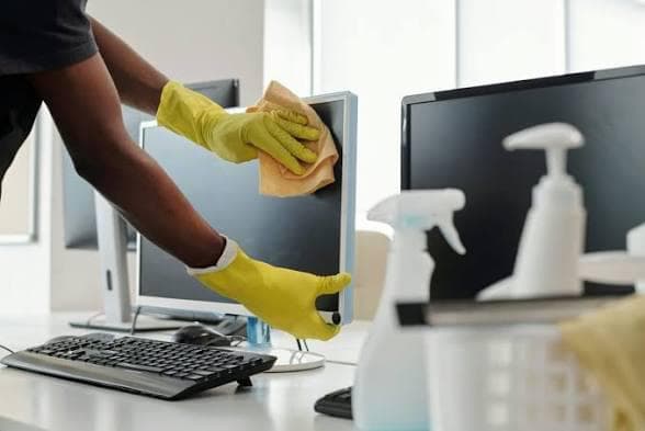 Cleaner wearing yellow rubber gloves carefully wiping a computer monitor with a microfibre cloth at a bright office desk