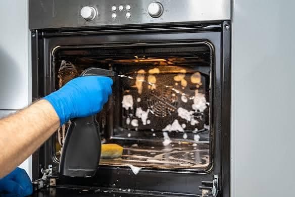 Cleaner spraying oven cleaner into a greasy built-in oven, with foam breaking down burnt-on residue on the back panel