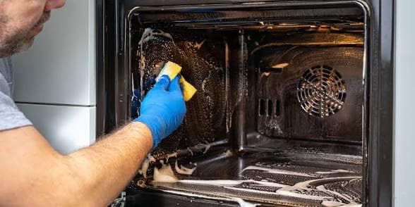 Professional cleaner wearing blue gloves scrubbing a greasy oven interior with a sponge and soapy foam