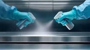 Two cleaners wearing blue gloves spraying disinfectant mist onto a stainless steel work surface during professional sanitisation