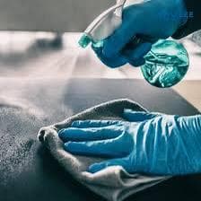 Close-up of gloved hands spraying disinfectant onto a dark countertop and wiping with a microfibre cloth during touch-point sanitisation