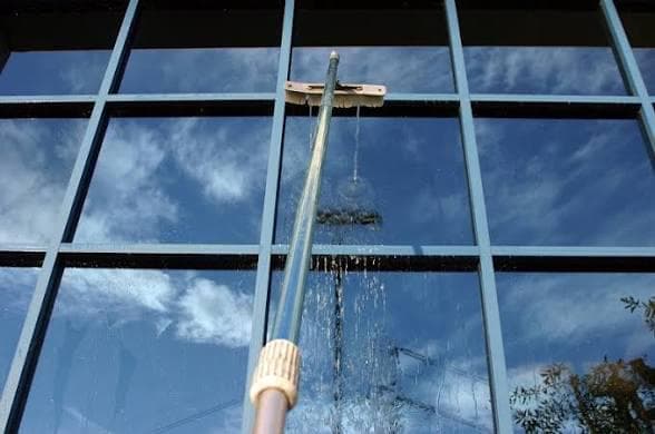 Professional window cleaner using a water-fed pole to wash exterior glass panes, with blue sky reflected in the windows